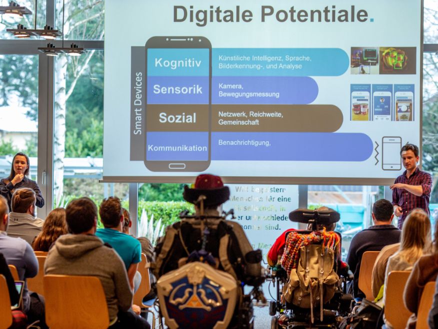 Presenter sharing research on digital accessibility potentials — cognitive, sensory, social, and communication dimensions — to a diverse seated audience including wheelchair users